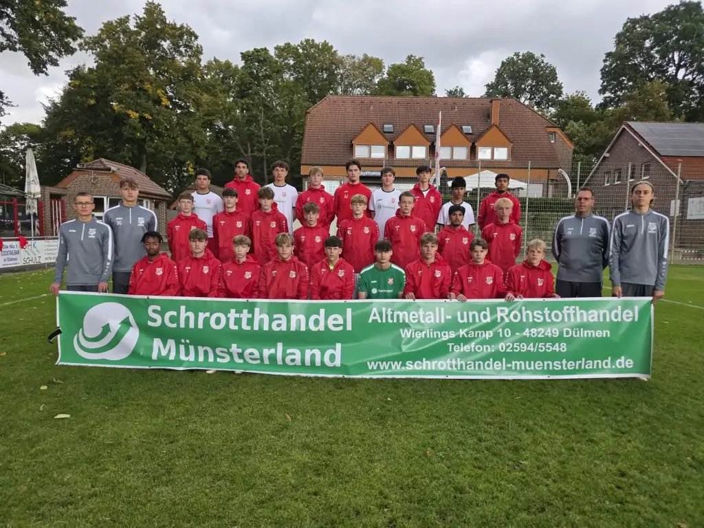 Jugendmannschaft in roten Trainingsjacken mit Trainern hinter dem Banner von Schrotthandel Münsterland auf dem Sportplatz